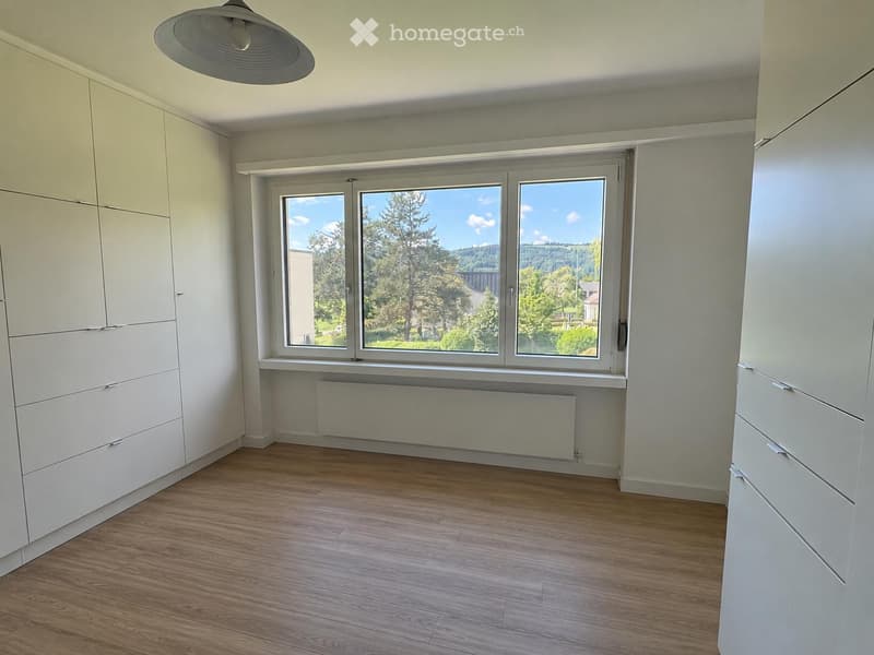 Empty room with wooden flooring, white walls, built-in wardrobes, large window with view, and radiator below the window.