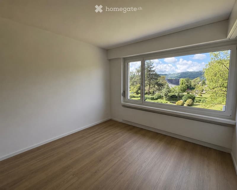 Empty room with wooden flooring, white walls, and a large window offering a view of a green landscape.