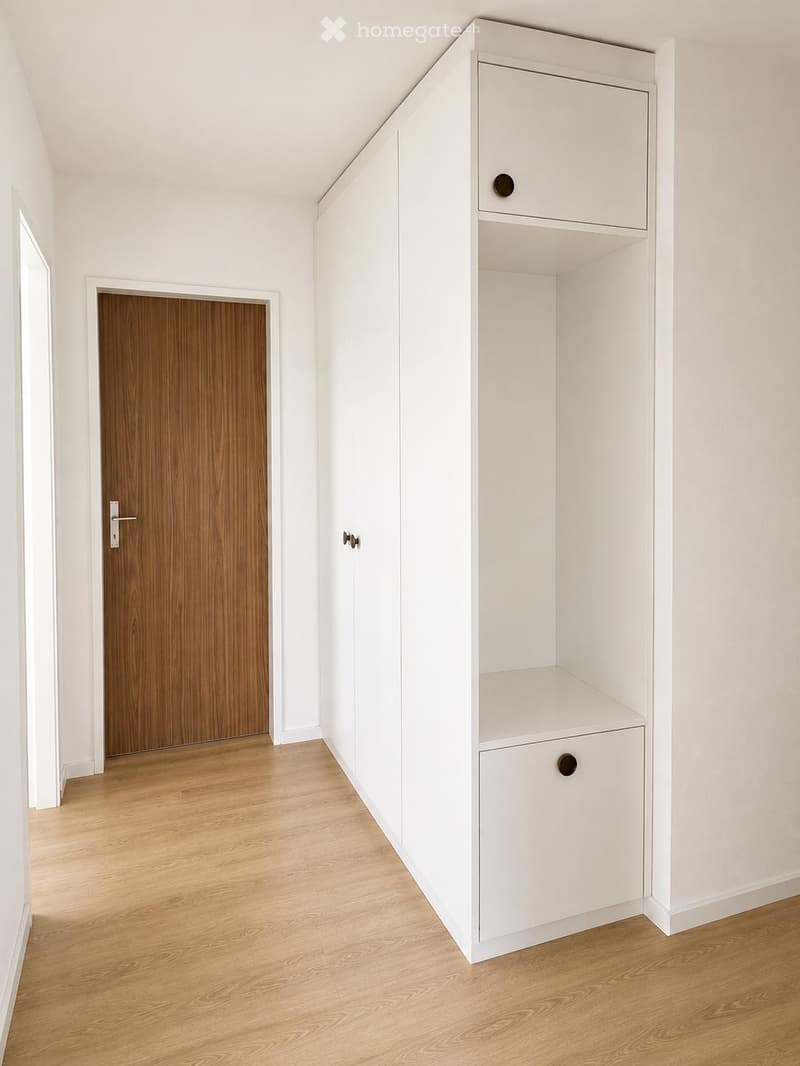 A clean, modern hallway with light wooden flooring, white walls, and a built-in white storage unit featuring shelves and a drawer. A wooden door is visible on the left side.