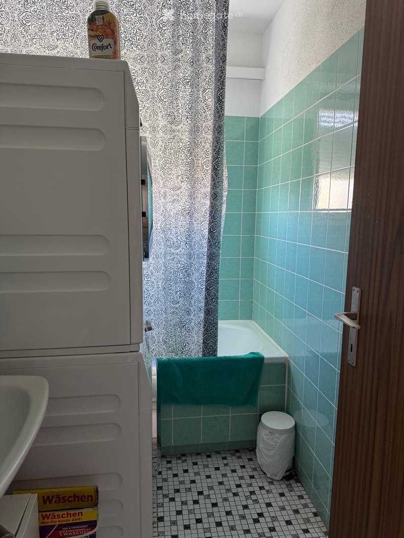 Small bathroom with green tiled walls, a bathtub with a towel draped over the edge, a shower curtain, and a toilet. The floor has a black and white checkered tile pattern.