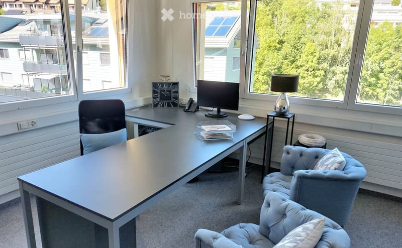 Office space with a large desk, computer screen, office chair, two armchairs, a lamp, a clock, and a telephone. Large windows offer a view of greenery and neighboring buildings.