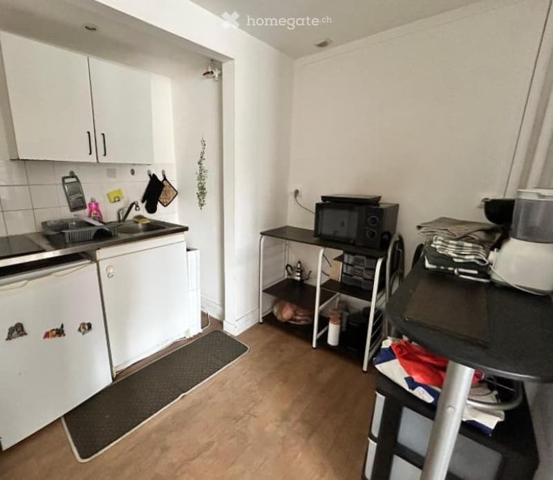 Compact kitchen with white cabinets, a sink, a dishwasher, a microwave, and a small table with a chair. The floor is wooden, and there are various kitchen items and utensils visible.