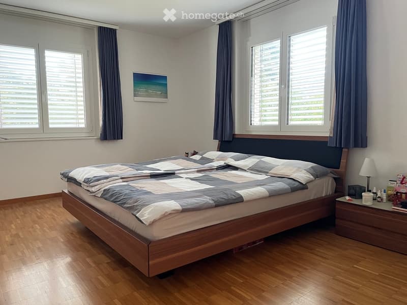 Well-lit bedroom with wooden flooring, large windows with blinds, a neatly made bed with a checkered duvet, a nightstand with a lamp, and a picture frame on the wall.