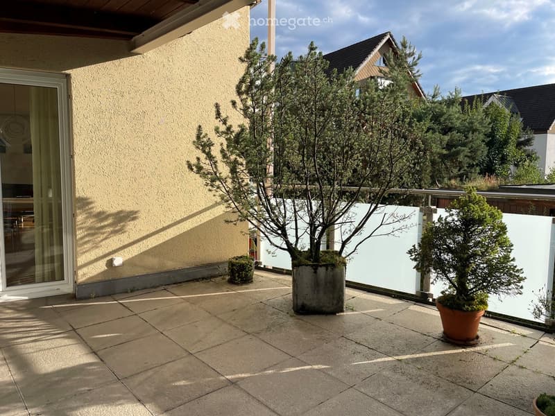 Balcony area with tiled flooring, potted plants, and a white privacy screen. The adjacent building has a light yellow facade and a large glass sliding door.