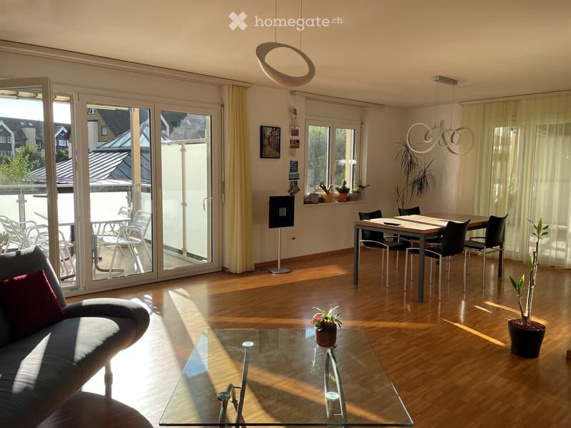 Bright living room with hardwood floors, large sliding glass doors leading to a balcony, a grey sofa with red pillow, a glass coffee table, a dining area with a table and chairs, and several potted plants.
