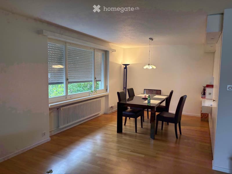 Dining room with wooden flooring, large window with blinds, dining table with chairs, a bottle, and a candle on the table, and a floor lamp near the window.