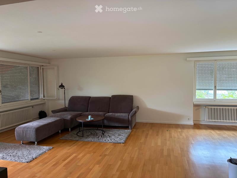 Modern living room with wooden flooring, large windows, grey sectional sofa, coffee table, ottoman, floor lamp, and mirror.