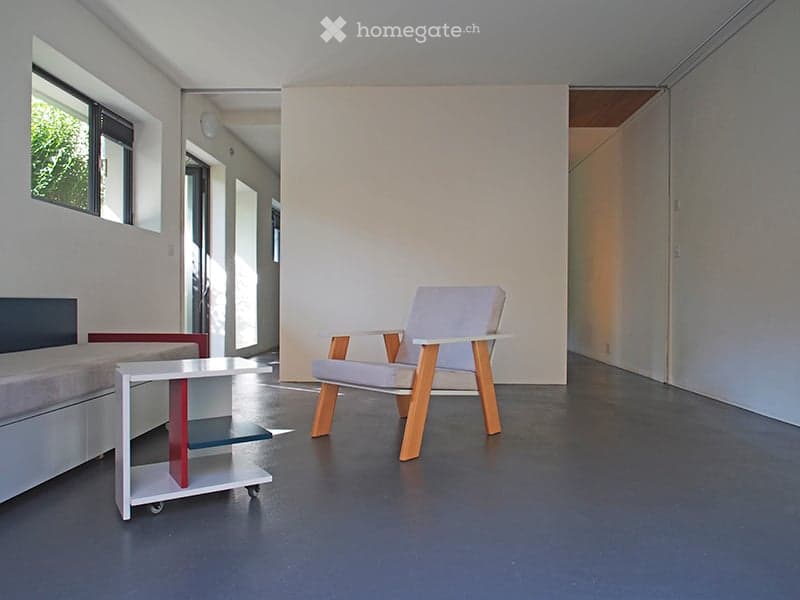 A modern, minimalist room with a grey sofa, a white wooden chair, and a small white table with red accents. The room features large windows and a clean, contemporary design.