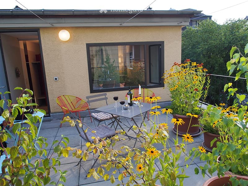 Balcony with a small table, chairs, and potted flowers, adjacent to a building with a large window and a lit wall light.