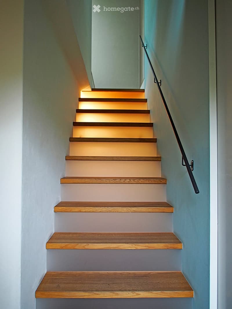 Modern staircase with wooden steps and a sleek black handrail, illuminated by warm ambient lighting.