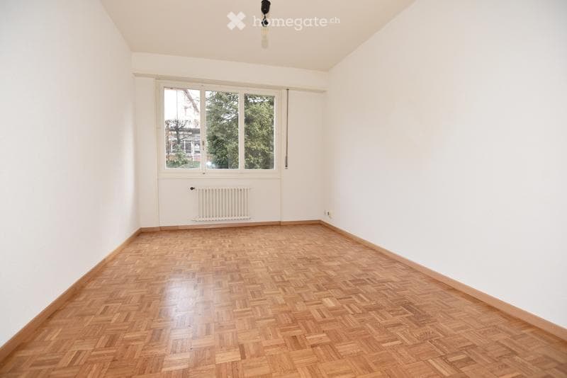Empty room with wooden parquet flooring, white walls, large window with view, radiator, and ceiling light fixture.