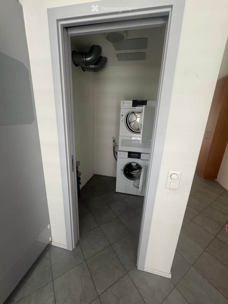 Small tiled room with a washing machine and dryer stacked vertically, white walls, and a door leading to the hallway.