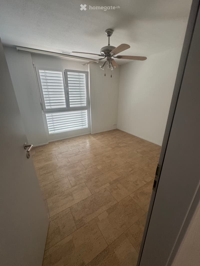 Empty room with wooden flooring, white walls, ceiling fan, and large window with blinds.