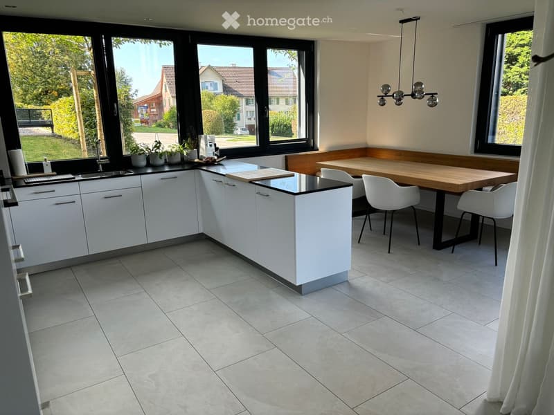 Modern kitchen with white cabinets, a central island, and a dining area with a wooden table and white chairs. Large windows provide natural light, and the space includes a sink, cutting board, and potted plants on the counter.
