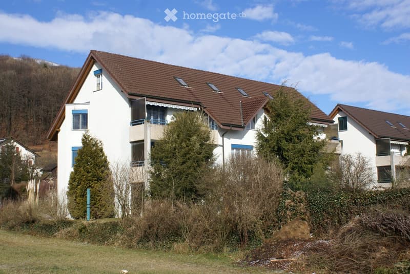 Two-story white house with brown tiled roof, multiple windows, and balconies surrounded by trees and shrubs.