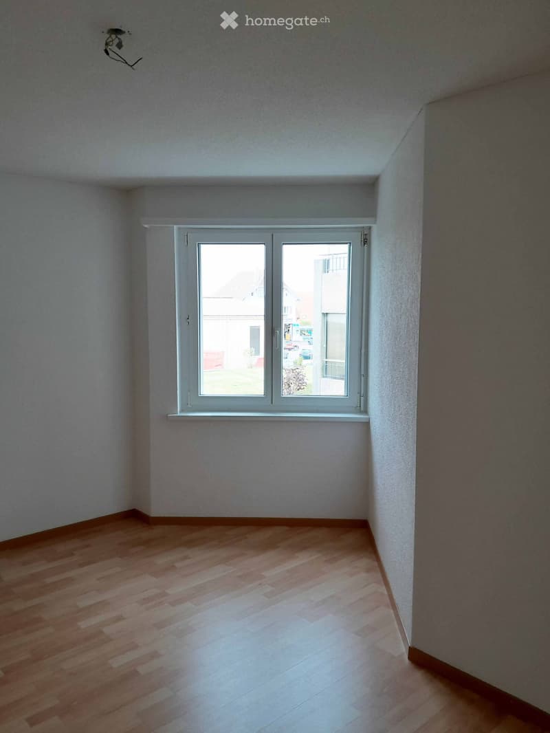 Empty room with wooden flooring and white walls, featuring a large window with white frame and a small electrical outlet on the wall.