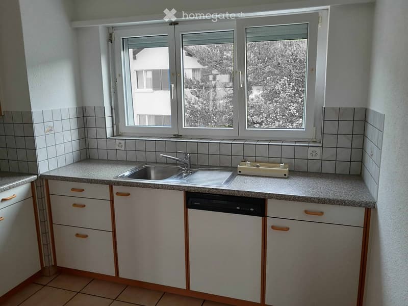 A compact kitchen with white tiled walls, granite countertops, a stainless steel sink, and a built-in dishwasher. The space features three windows offering a view of the outside.