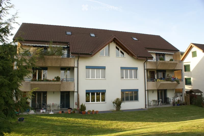 Two-story house with a tiled roof, multiple windows, balconies, and a well-maintained lawn with potted plants and outdoor furniture.