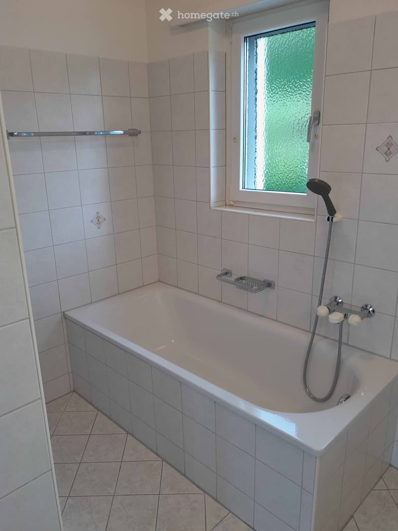 Clean bathroom with a bathtub, tiled walls, a window with frosted glass, and a showerhead attached to the tub.