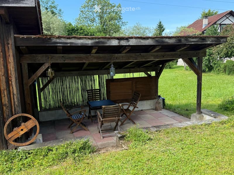 Wooden outdoor seating area with a table and chairs under a covered wooden structure, surrounded by green grass and trees.