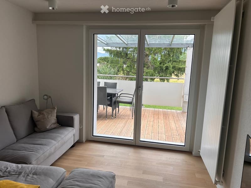 Living room with grey sofa, ottoman, and sliding glass doors leading to a balcony with outdoor furniture.