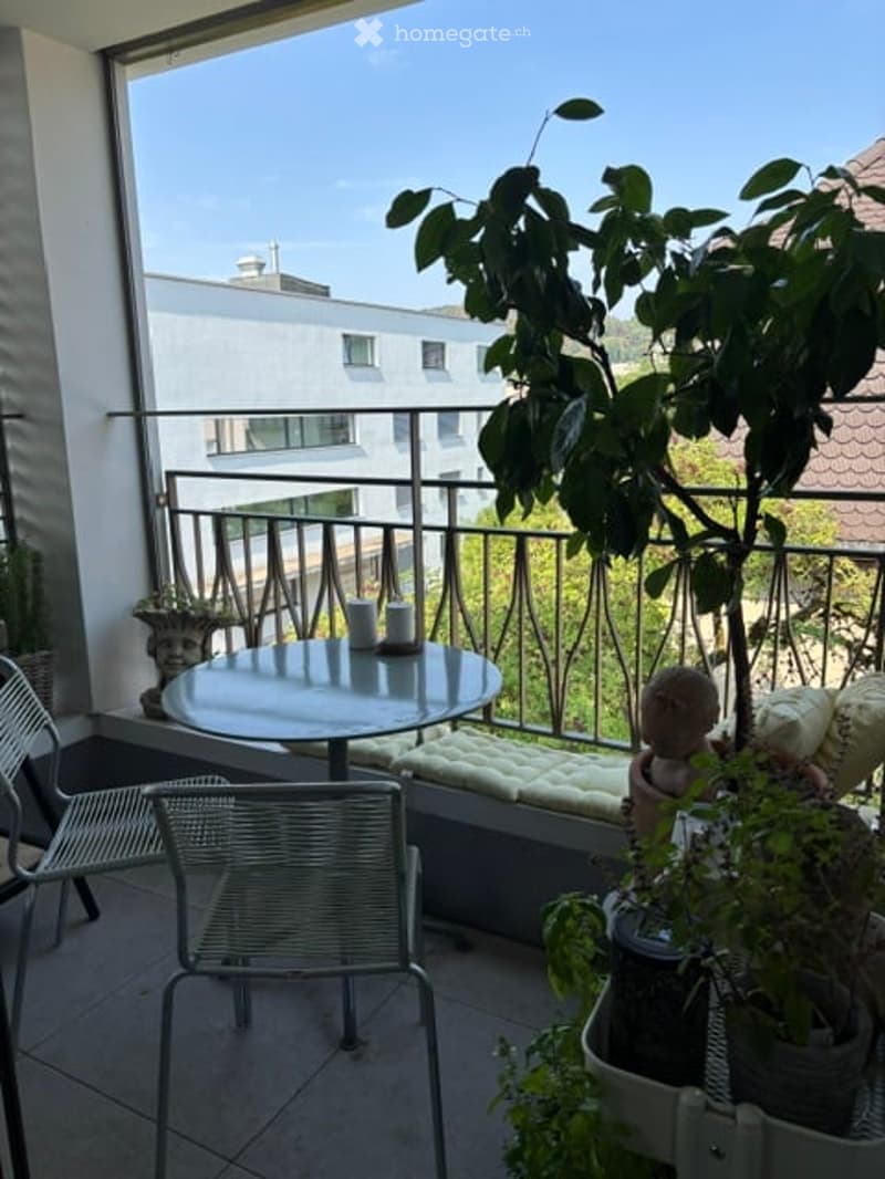 Balcony with a round table, chairs, and potted plants, overlooking a cityscape with buildings and greenery.