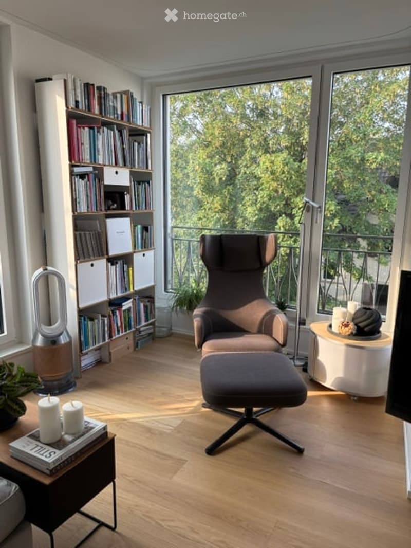 Modern living room with large windows offering a view of greenery, featuring a brown sofa, ottoman, bookshelf, small table with candles, and a sleek lamp.