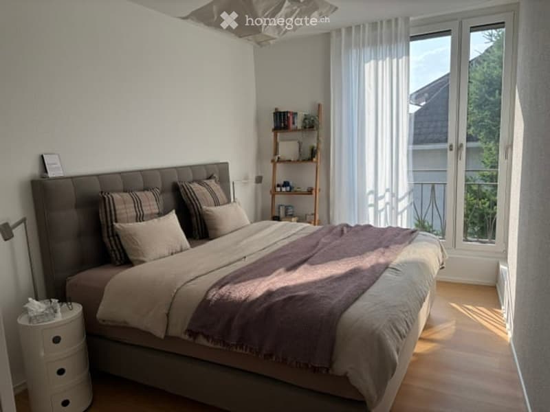 Modern bedroom with a large bed, nightstand, shelf, and large windows offering a view of greenery.