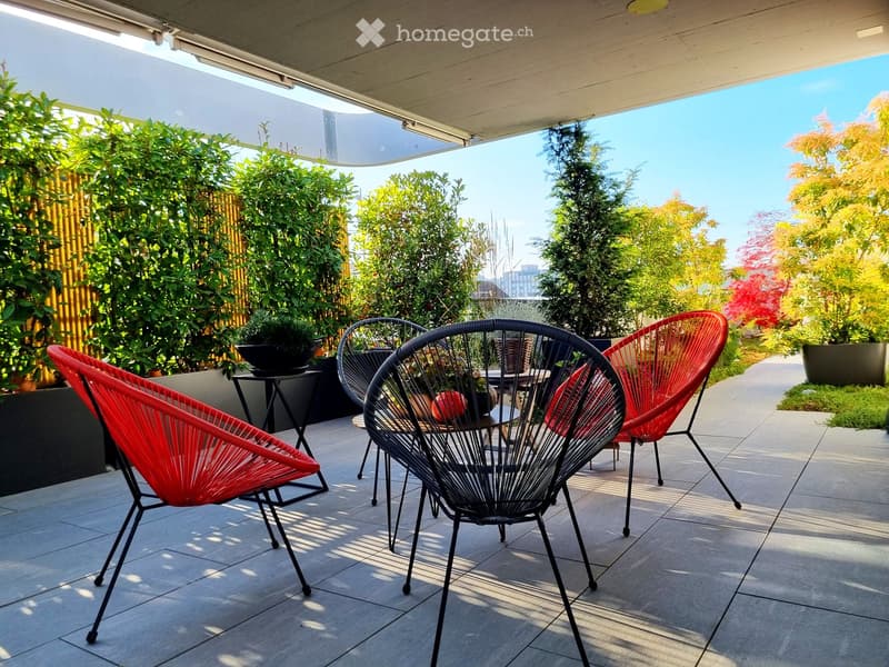 A modern outdoor patio with red and black wire chairs, a small table, and lush greenery in the background.
