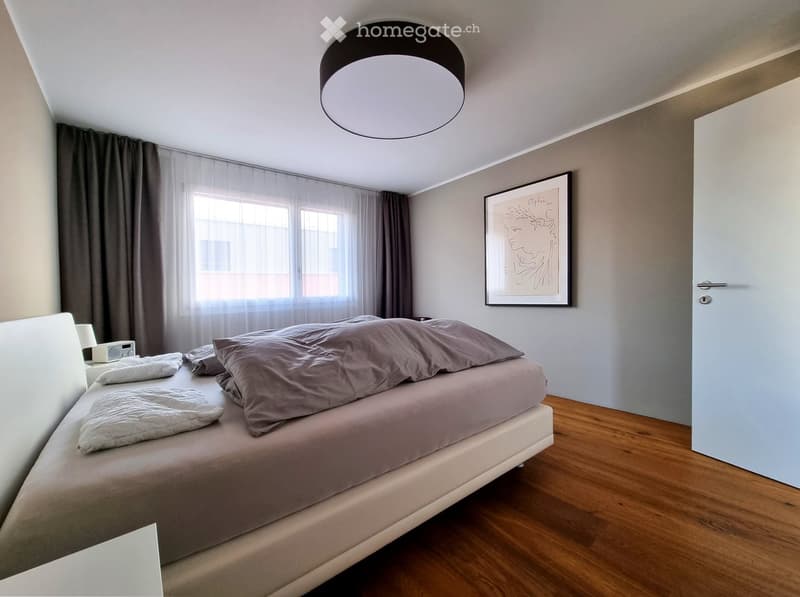 Modern bedroom with a large bed, pillows, nightstand, lamp, and a framed picture on the wall. The room features wooden flooring and large windows with curtains.