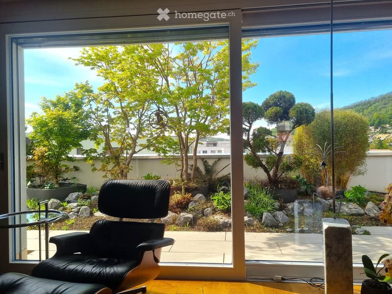 A modern room with large windows offering a scenic view of a landscaped garden. The room features a black leather chair and a glass table.