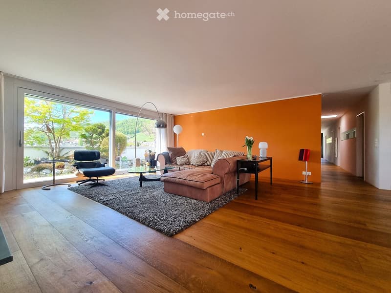 Modern living room with wooden flooring, large glass doors leading to a garden view, orange accent wall, and contemporary furniture including a sofa, ottoman, armchair, coffee table, and side table.