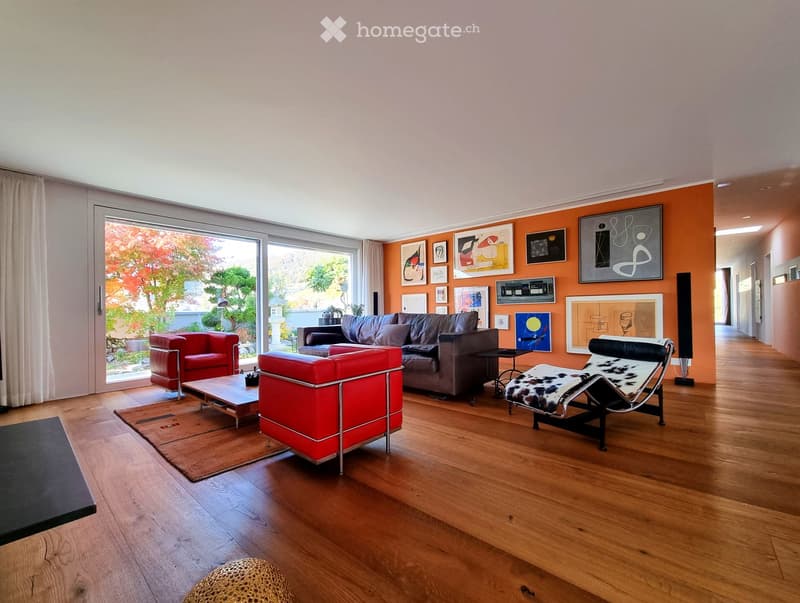 Modern living room with wooden flooring, large windows, and contemporary furniture including a sofa, chairs, coffee table, and artworks on the wall.