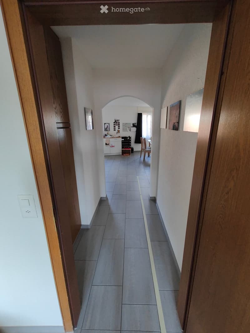 A long, narrow hallway with tiled flooring and white walls, featuring an arched opening leading to another room with visible furniture and decor.
