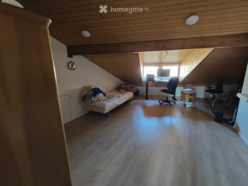 Loft-style room with wooden floors and slanted ceilings, featuring a bed, desk with laptop, office chair, bicycle, and storage boxes.