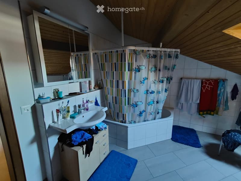 Bathroom with a sink, mirror, bathtub with a colorful fish-themed shower curtain, towels, and a blue bath mat on the floor.