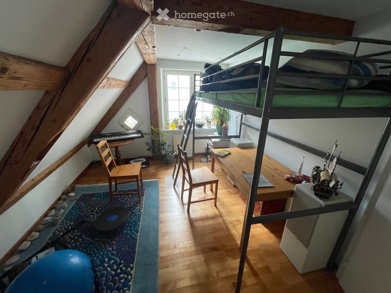 Cozy attic room with wooden floors, slanted ceilings, bunk bed, desk, chairs, and small table with keyboard. Features a blue rug, plants, and a toy on the desk.