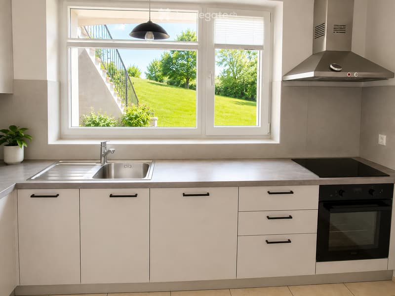 Modern kitchen with white cabinets, stainless steel sink, oven, induction cooktop, and range hood. Large windows offer a view of a green lawn and garden.