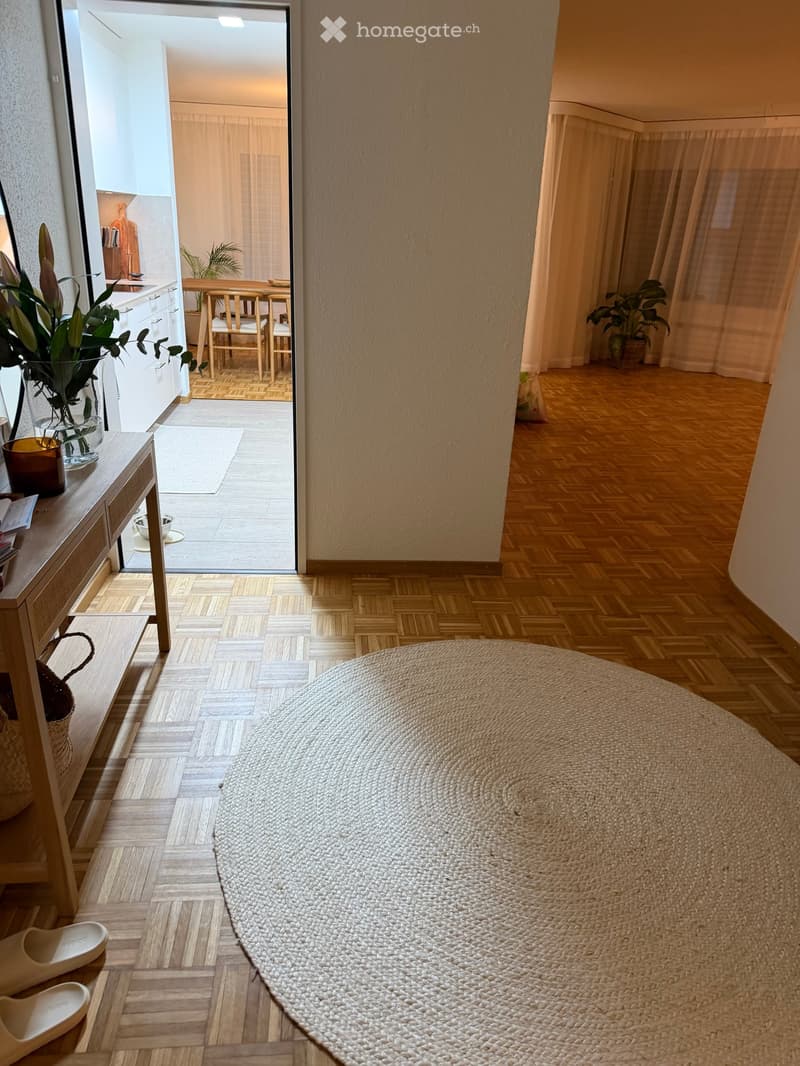 entryway with wooden floor, round woven rug, wall-mounted mirror, table with vase, shoe rack, doorway leading to kitchen