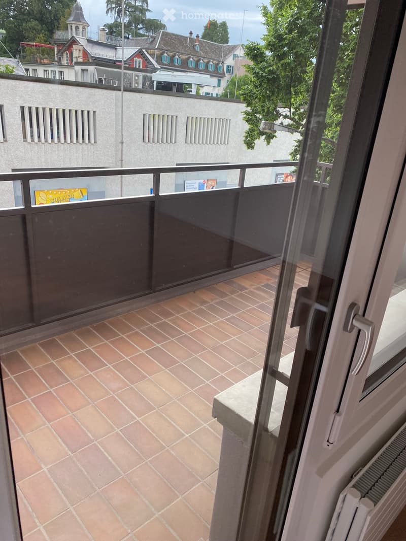 Balcony with tiled floor, glass railing, and view of surrounding buildings and greenery.