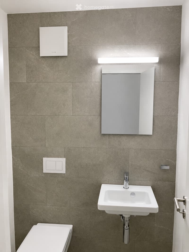 Modern bathroom with a white sink, chrome faucet, large mirror, and toilet. Grey tiled walls and built-in lighting above the mirror.