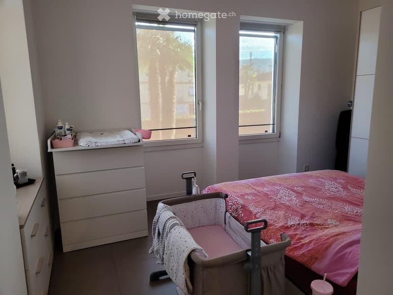 A small bedroom with a double bed, a baby crib, a nightstand, and a drawer unit. The room has two windows with blinds and a shelf above the nightstand holding various items.
