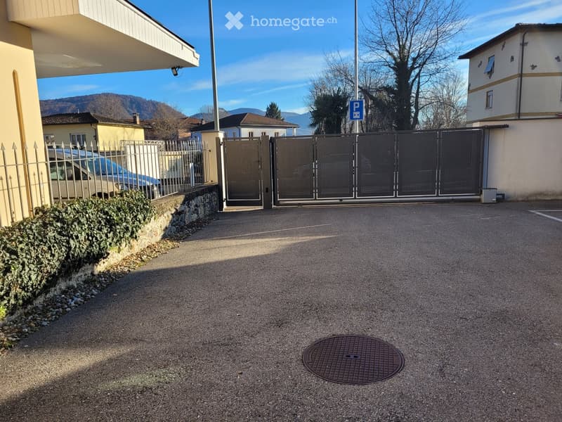 Driveway with gravel surface, metal gate, parked car, stone wall, and surrounding buildings.