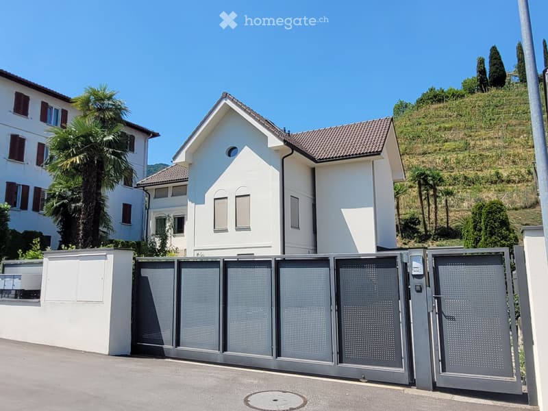 A modern two-story white house with a tiled roof, surrounded by greenery and a gated entrance.