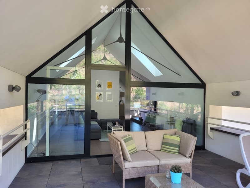Modern, open-plan living space with large glass walls and a high, slanted ceiling. Features a wicker sofa with striped cushions, a small coffee table, a potted plant, and an ottoman. The space is bright and airy with a view of the outdoor area.