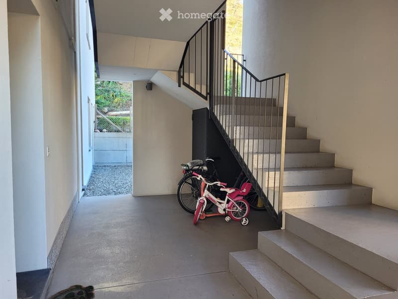A modern, open corridor with a staircase leading upwards, featuring a metal railing and concrete steps. A small child's bicycle is parked near the base of the stairs, and the area has a clean, minimalist design with light-colored walls and a tiled floor.