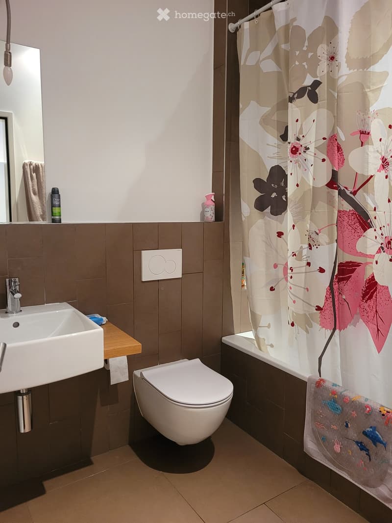 Modern bathroom with a white sink, toilet, bathtub, and a floral shower curtain. Includes a mirror, towel, and small decorative items on the counter.