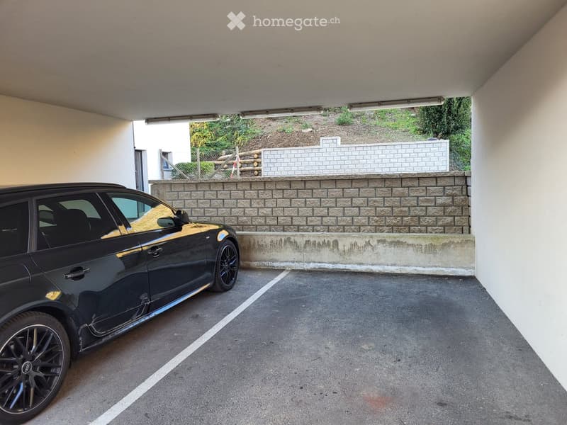 Underground parking area with a black car parked inside, featuring concrete walls, a brick wall in the background, and a raised ground floor visible through an opening.