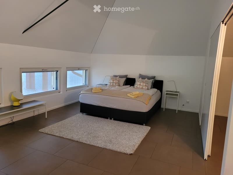 Modern bedroom with a large bed, nightstand, table lamp, bench, and a white rug on the floor.