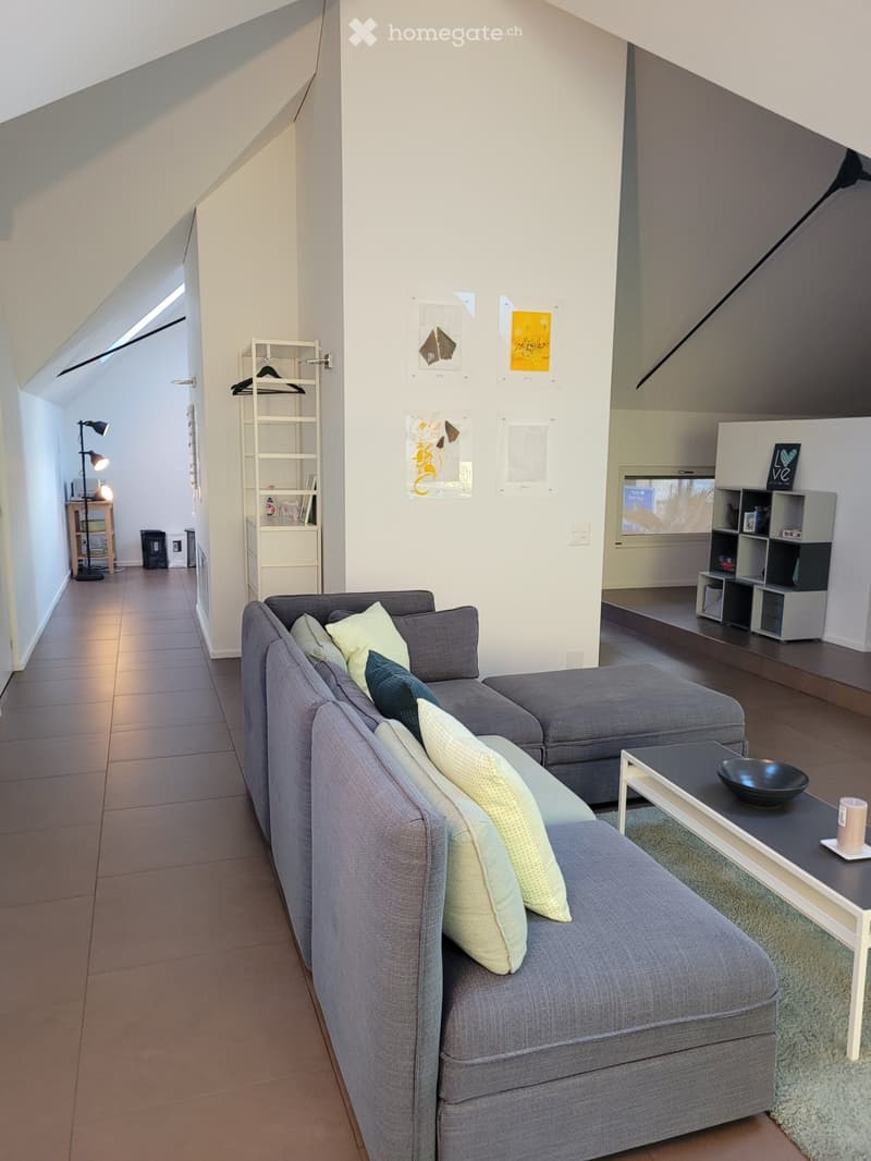 Modern living room with grey sectional sofa, coffee table, shelves, and a lamp. The room features a slanted ceiling and large windows allowing natural light.